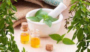 Two glass bottles of aromatherapy essential oils with cork stoppers with green herbs in a white mortar bowl with pestle for an ingredient article about herbalists formulating natural skincare products