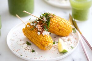 Two Elote Mexican Street corn on white plate with cotija cheese, tajin, lime, and cilantro for trend article about the flavor